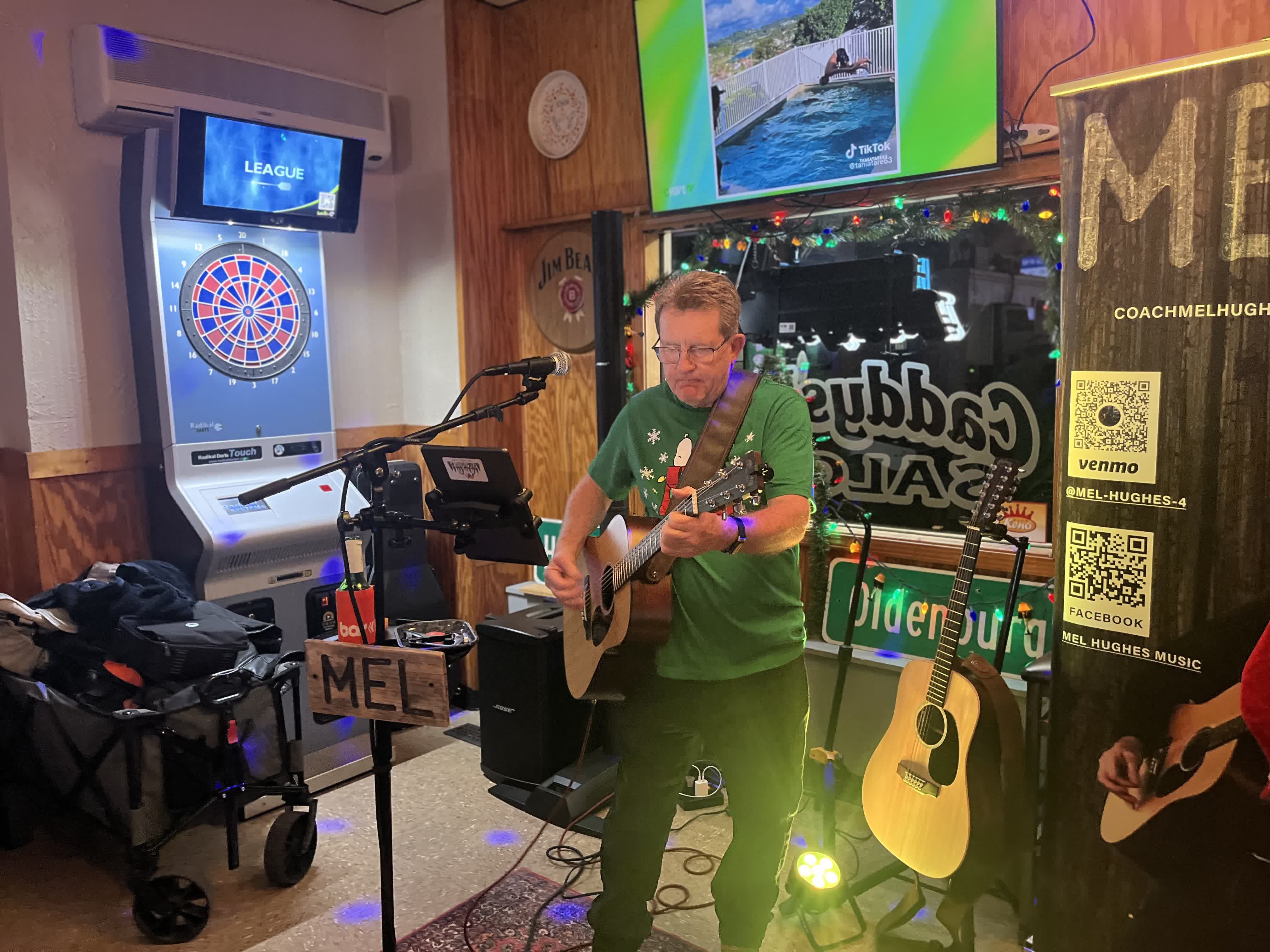 Mel Hughes performing in a bar with a dartboard and his banner behind him