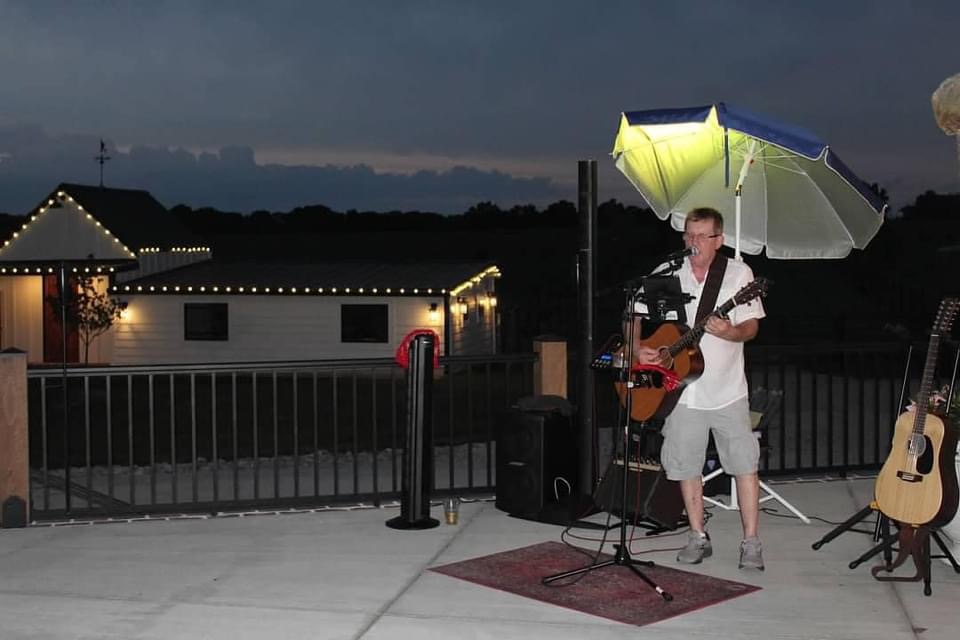 Mel Hughes performing at dusk on a patio, guitar in hand under string lights