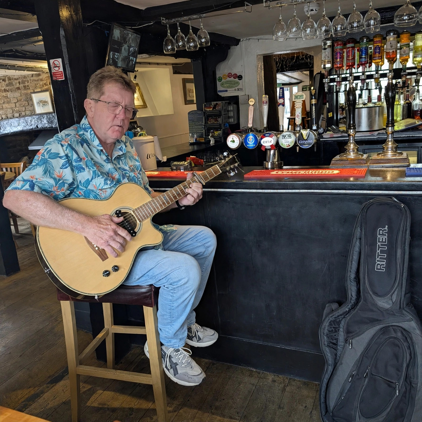 Mel Hughes sitting on a stool at a pub bar in Hythe, England, playing his acoustic guitar
