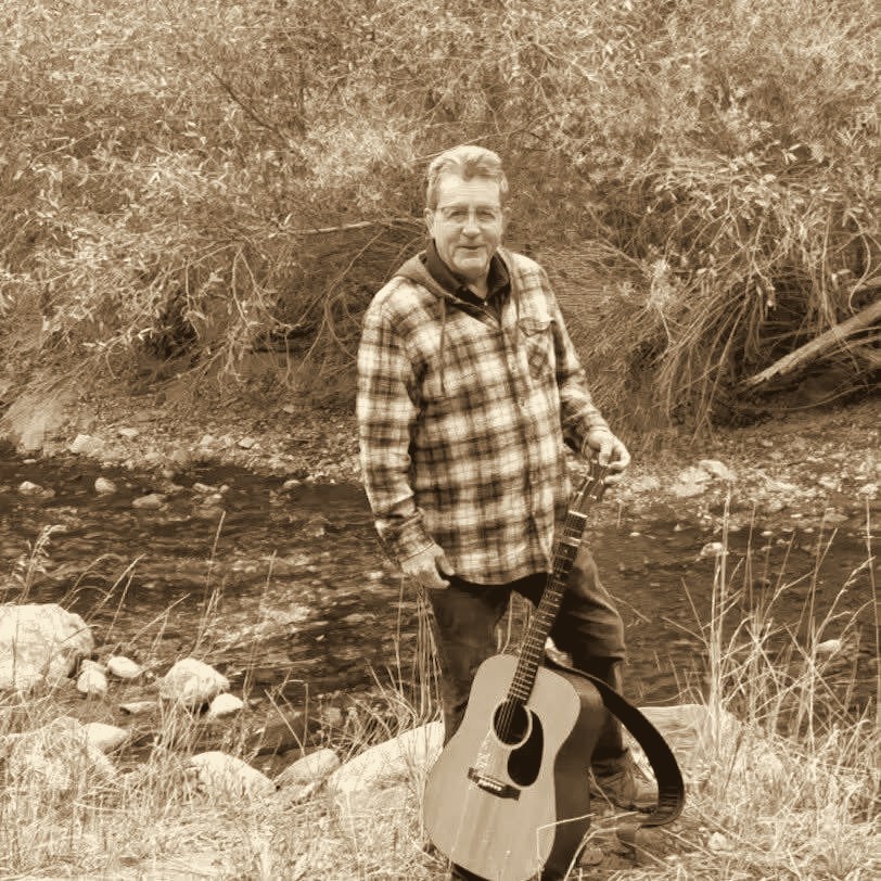 Mel Hughes standing on a creek bank in a plaid shirt, holding his acoustic guitar